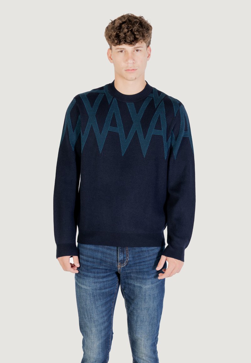 Jeune homme portant un pull bleu marine avec un motif en zigzag bleu sur la poitrine et les épaules, associé à un jean bleu délavé, se tenant devant un fond uni.
