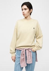 Νεαρός με κοντά σκούρα μαλλιά, φοράει μπεζ φούτερ, ριγέ μπλε τζιν και ροζ πουκάμισο δεμένο στη μέση, κοιτάζει αριστερά με το χέρι στην τσέπη.