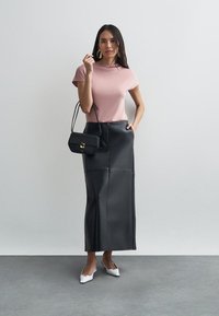 Różowy top z krótkim rękawem w połączeniu z czarną, skórzaną, długą spódnicą. Modelka trzyma małą czarną torebkę z złotym akcentem. Białe szpilki z wąskim noskiem.