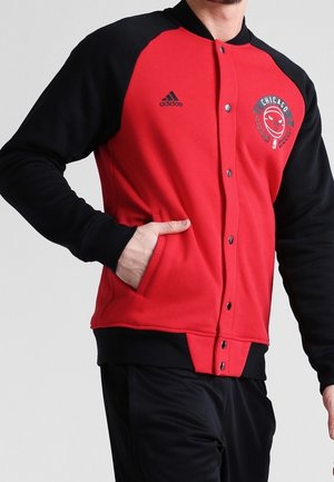 Person, die eine rot-schwarze Chicago Basketball Adidas-Jacke trägt, mit der Hand in der Tasche, sichtbar von den Schultern bis zu den Hüften.