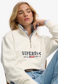 Kobieta o blond włosach nosi kremową bluzę Superdry z ćwierćzamkiem na zamek błyskawiczny, pod którą ma niebieską koszulę w paski z kołnierzykiem, siedząc ze skrzyżowanymi nogami.