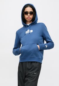 Μπλε φούτερ με κουκούλα και τσέπη μπροστά, με λευκό λογότυπο Nike. Συνδυασμένο με μαύρο παντελόνι που έχει μικρό λογότυπο Nike.