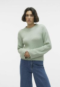 Pulover ribbed de un verde deschis, cu guler rotund și mâneci lungi, purtat împreună cu blugi din denim albastru. Materialul pare moale și texturat.