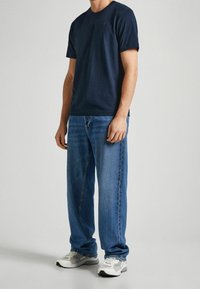 Hombre vistiendo una camiseta marina, jeans azules holgados y zapatillas blancas, de pie contra un fondo claro y liso.