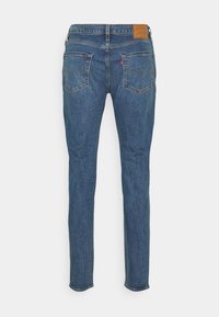 Vue arrière d'un jean en denim bleu avec deux poches arrière, un patch logo en cuir marron sur la ceinture et des jambes droites et fines.