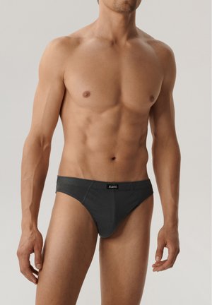 Umięśniony mężczyzna w ciemnoszarych majtkach z napisem „Atlantic”, stoi na jednolitym jasnym tle, widoczne tułów i górna część nóg.