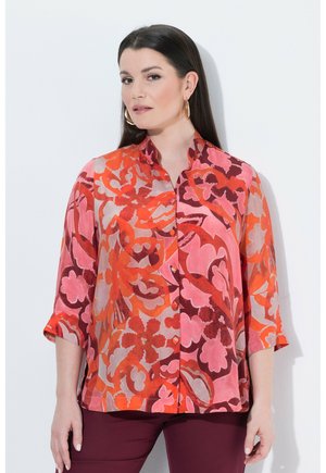 Frau mit langen dunklen Haaren trägt ein lockeres, mehrfarbiges Blumenhemd in Rot-, Pink- und Orangetönen und große goldene Creolen.