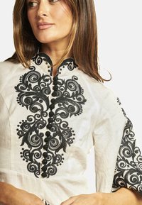Blusa ricamata in lino beige, con intricati motivi floreali neri, colletto alto con bottoni e maniche corte.