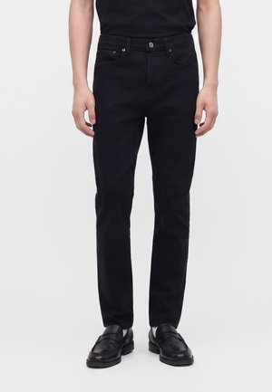 Homme portant un jean noir droit, une ceinture noire, des mocassins noirs et un t-shirt noir sur un fond uni.