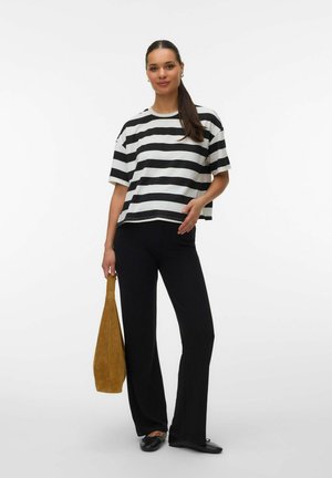 Naine pikkade tumedate juustega, kandes musta-valge triibulist särki ja musti pükse, hoides pruuni käekotti, seisab valgel taustal.