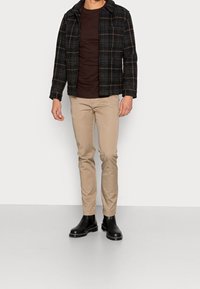 Herrenoutfit mit einer schwarz-braun karierten Jacke, einem braunen T-Shirt, hellbeigen Hosen und schwarzen Stiefeletten. Glatter Stoff, maßgeschneiderte Passform.