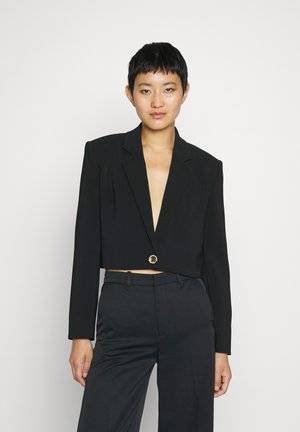 Blazer noir court en tissu lisse, avec des revers crantés, des manches longues et une fermeture à bouton circulaire à l'avant.