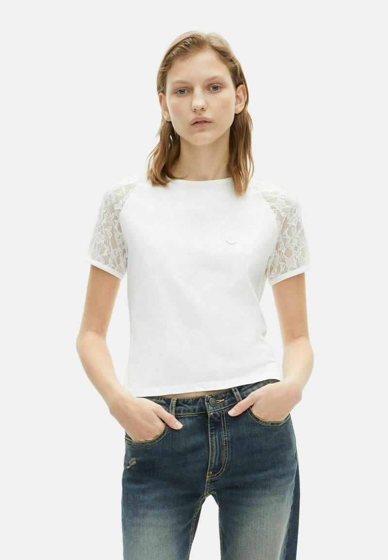 Witte t-shirt met korte mouwen en kantaccent op de mouwen. Heeft een ronde hals en een aansluitend ontwerp, gecombineerd met blauwe spijkerbroek.
