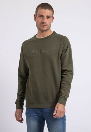 Homme portant un sweat-shirt vert olive avec le texte "Ritchie Jeans Co." et un jean bleu, debout devant un fond blanc uni.