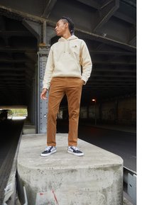 Beige hettegenser, brune kordfløyelsbukser og svart-hvite rutete sneakers brukt av en person som står på en betongblokk under en motorvei.