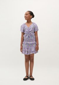 Lila Blumenkleid mit kurzen Ärmeln, gerafftem Vorderteil und gestuftem, rüschengeschmücktem Rock. Kombiniert mit schwarzen Ballerinas.