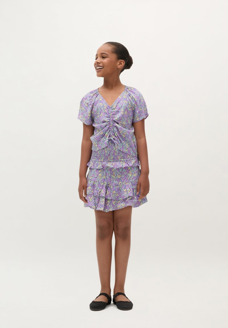 Lila Blumenkleid mit kurzen Ärmeln, gerafftem Vorderteil und gestuftem, rüschengeschmücktem Rock. Kombiniert mit schwarzen Ballerinas.