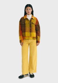 Chaqueta de cuadros en colores naranja, amarillo y marrón con dos bolsillos en el pecho y botones, combinada con pantalones de pana amarillos de pierna ancha. Zapatillas negras.