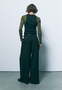 Črne široke hlače v kombinaciji s prilegajočo, temno majico z olivno zelenimi rebrastimi rokavi. Gladka tkanina z subtilno teksturo in čistimi linijami.
