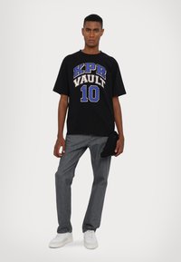 Schwarzes Baumwoll-T-Shirt mit großen blauen, weißen und beigen Grafiken, auf denen "KPR VAULT 10" steht, kombiniert mit grauen Jeans und weißen Sneakers.