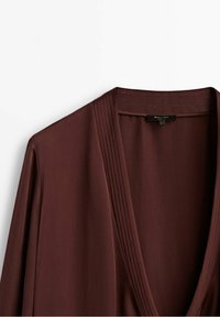 Ruskea kääreblouse syvällä V-kaula-aukolla, jossa on hienovaraisia teksturoituja poimuja kaula-aukon ympärillä ja sileä kangas hienostuneen ilmeen saavuttamiseksi.
