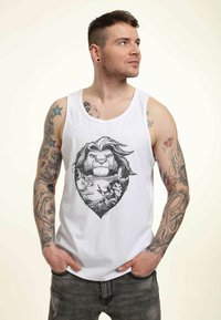 Hvid tanktop med et sort grafisk design af et løves ansigt sammen med landskabs- og dyreillustrationer. Modellen har tatoveringer og grå jeans.