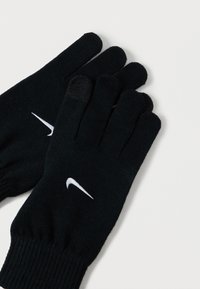 Un paio di guanti in maglia nera con i loghi Swoosh bianchi di Nike, un guanto mostra la punta compatibile con il touchscreen, esposti su uno sfondo bianco.