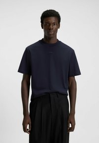 Mladý černoch stojící v námořnickém modrém tričku s krátkým rukávem značky Hugo a černých plisovaných kalhotách na pozadí jedné světle barvy.