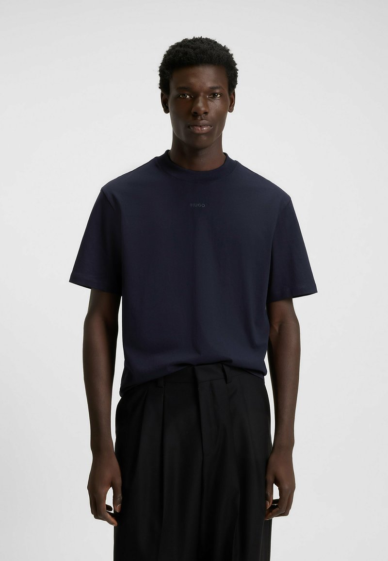 Mladý černoch stojící v námořnickém modrém tričku s krátkým rukávem značky Hugo a černých plisovaných kalhotách na pozadí jedné světle barvy.