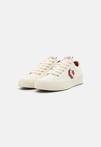 Canvas low-top sneakers in cream, featuring maroon accents, a star logo, and textured rubber sole with a diamond pattern.