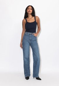 Frau mit langen schwarzen Haaren, die ein schwarzes Tanktop, hoch taillierte blaue Jeans und schwarze spitze Schuhe trägt, steht vor einem weißen Hintergrund.