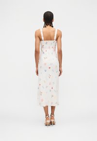 Γυναίκα με πλεξούδες που φοράει λευκό midi φόρεμα με πολύχρωμα σχέδια με θαλασσινά θέματα και λευκά ψηλοτάκουνα σανδάλια, στέκεται γυρισμένη με την πλάτη.