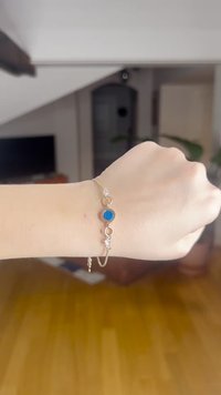 Pulseira de ouro com uma pedra circular azul como destaque, detalhes de contas transparentes e uma corrente delicada. Usada num pulso contra um chão de madeira.