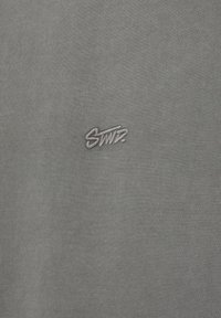Tissu gris avec petit logo brodé "STMP." en fil gris assorti centré sur la surface.