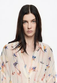 Blusa ligera y transparente con patrones florales multicolores sobre un fondo beige, con detalles de botones y un corte relajado.