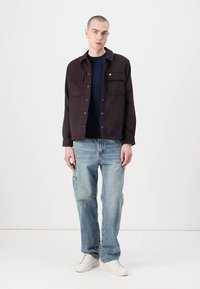 Mørkebrun skjorte med knapper og to brystlommer, båret over en navy skjorte og parret med lyseblå slidte jeans og hvide sneakers.