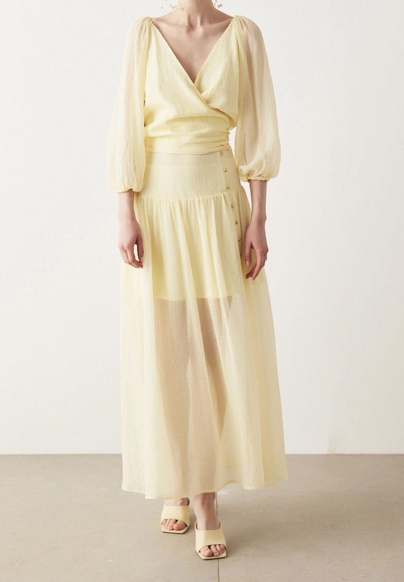 Keltainen kietaisumekko, jossa on ilmapallon hihat, teksturoitu kangas ja nilkkapituiset helmat. Sisältää sivunapit ja läpikuultavan päällikankaan. Yhdistetty keltaisiin avokärkisiin kenkiin.