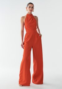 Oranžový overal s dizajnom na halterneck, širokými nohami, textúrovanou tkaninou a priliehavým pásom. Štylizovaný s veľkými okrúhlymi náušnicami.