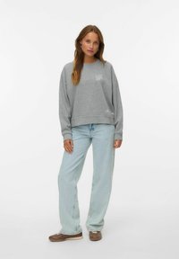 Felpa grigia con colletto a girocollo e spalle basse. Presenta un piccolo logo LA e una scritta. Abbinata a jeans azzurri chiari e dalla vestibilità ampia.