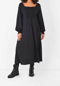 Robe noire à manches longues avec un décolleté carré, corsage smocké et jupe fluide, associée à des bottes de combat noires à lacets.