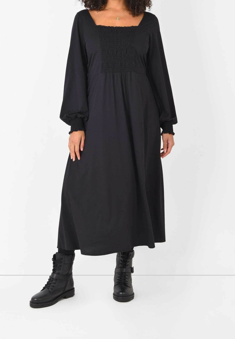 Robe noire à manches longues avec un décolleté carré, corsage smocké et jupe fluide, associée à des bottes de combat noires à lacets.