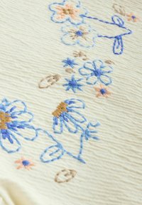 Tissu crème orné de fleurs brodées en bleu, pêche et brun. Le design comprend divers motifs floraux et des détails de couture intriqués.