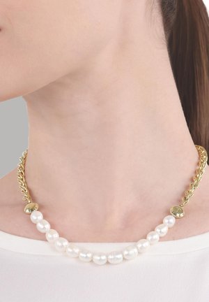 Ketting met grote witte parels en een dikke gouden ketting, gedragen om de hals van een vrouw tegen een lichte topachtergrond.