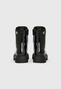 Bottines en cuir verni noir avec une semelle épaisse, détail de fermeture éclair et accent logo à l'arrière. Texture lisse et design épuré.
