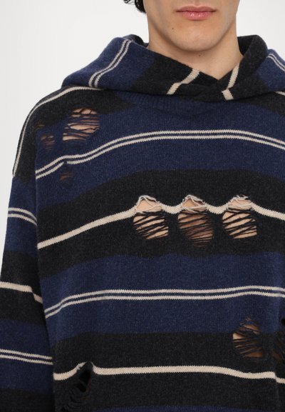 Pull en bleu et noir rayé avec capuche, présentant plusieurs trous usés et des rayures blanches en accent. Texture en tricot douce visible.