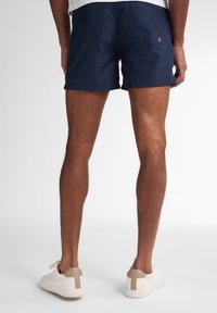 Pantaloni scurți de baie navy cu o croială relaxată, confecționați dintr-un material ușor. Au un mic detaliu cu logo roșu și un tiv plat. Purtați cu adidași albi.