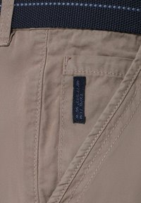 Beige Hosen aus glattem Stoff mit Steppakzenten. Ein dunkles Etikett ist teilweise sichtbar und zeigt Größen- und Markeninformationen.