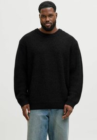 Mann mit Bart, der einen schwarzen Strickpullover und hellblaue Jeans trägt, steht vor einem schlichten, hellen Hintergrund.