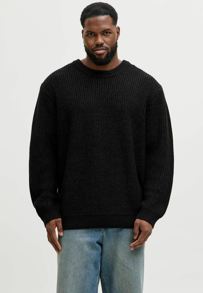 Mann mit Bart, der einen schwarzen Strickpullover und hellblaue Jeans tr�ägt, steht vor einem schlichten, hellen Hintergrund.