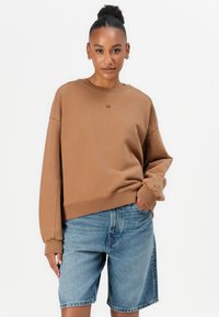 Ruskea fleece-sweatshirt, jossa on leveät hihat ja laskeutuvat olkapäät. Yhdistetty vaaleansinisiin farkkushortseihin, joissa on klassinen leikkaus ja etutaskut.